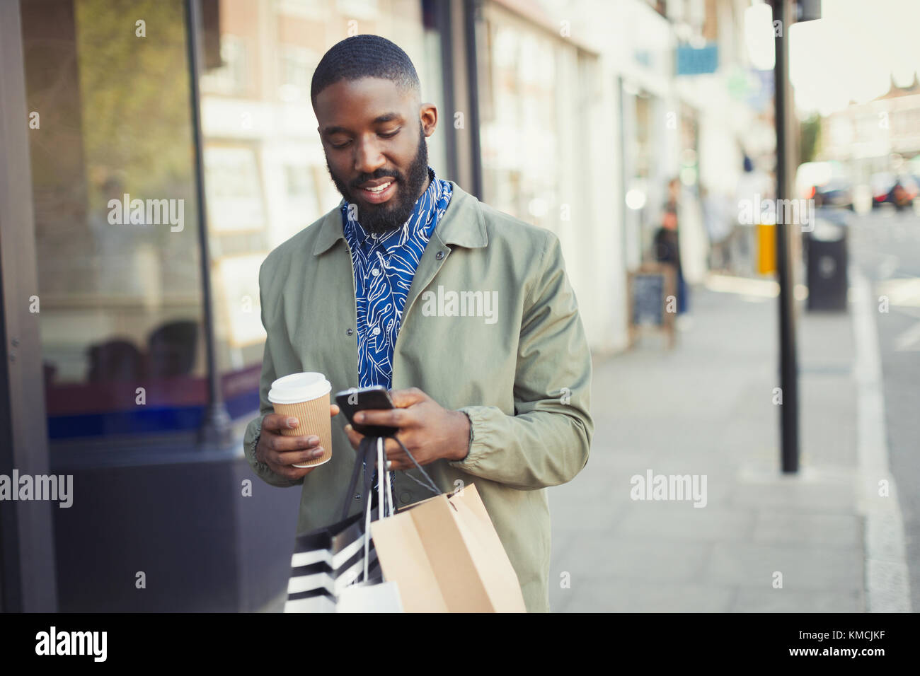 Giovane uomo con caffè e borse da shopping testando con cella telefono sul marciapiede urbano Foto Stock