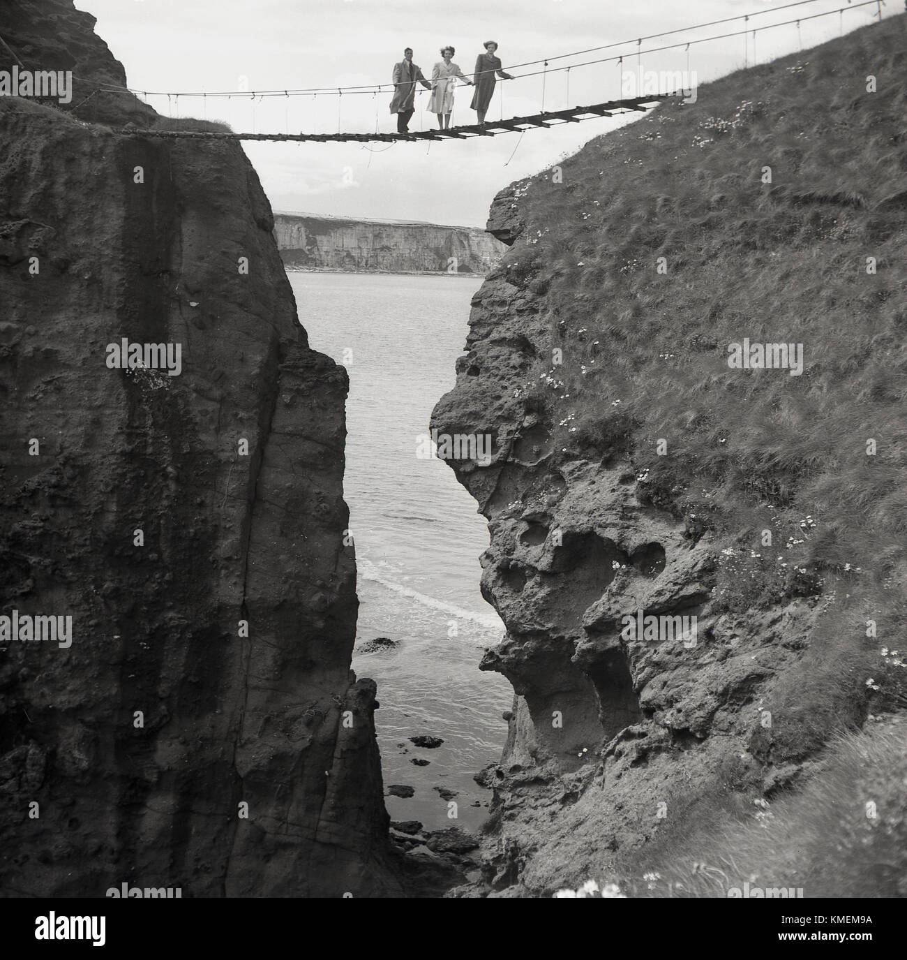 Degli anni Cinquanta, storico, un uomo e due donne posano per una foto in piedi sul famoso Carrick-a-Rede ponte di corde, Co Antrim, Irlanda del Nord. Il ponte di corde è quasi 100ft alta che collega la terraferma con la piccola isola di Carrickarede ed è stato definito per la prima volta nel 1755 dal salmone locale di pescatori. Foto Stock