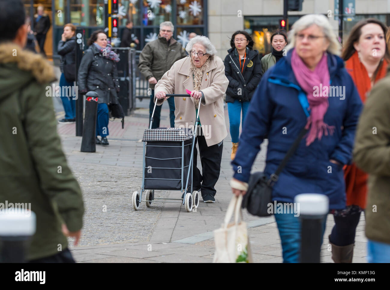 Donna anziana a camminare in una città frenetica in negozi con un carrello di shopping come parte di un camminatore gommati, NEL REGNO UNITO. Foto Stock
