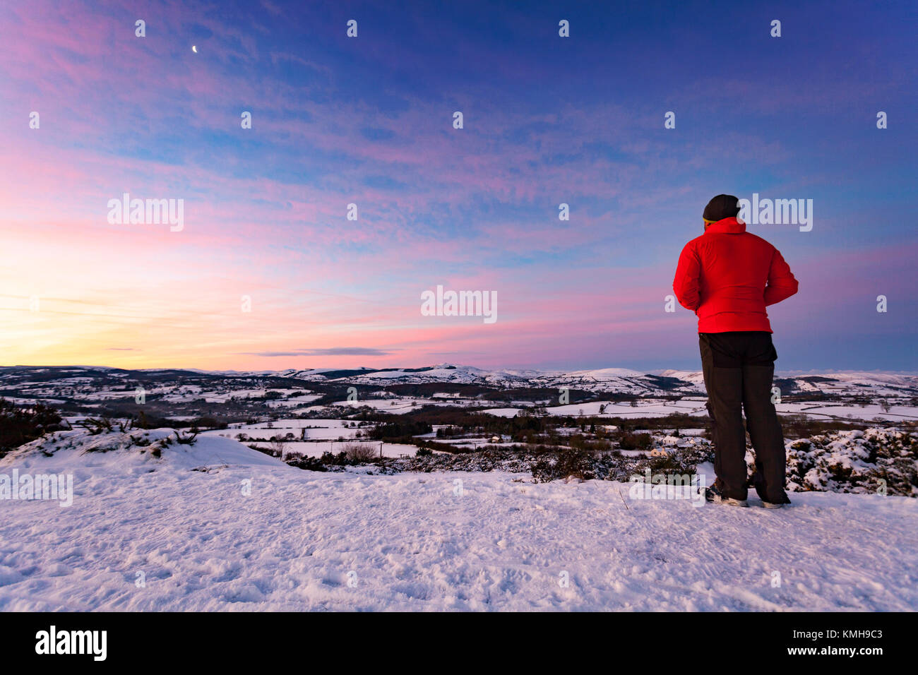 Un camminatore di montagna che si affaccia su paesaggi innevati della gamma Clwydian e Famau Moel durante un inverno sunrise dal piede delle colline di Moel y Gaer hill fort vicino Halkyn, Flintshire, Wales, Regno Unito Foto Stock