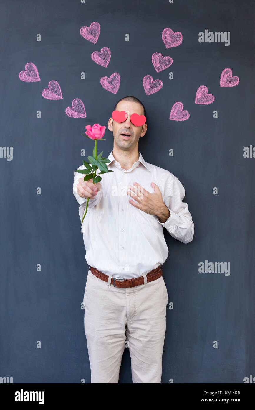 Singolo adulto uomo bianco con gli occhi a forma di cuore che indossa una camicia bianca e in piedi di fronte a una lavagna con dipinto di cuori tenendo una rosa. Foto Stock