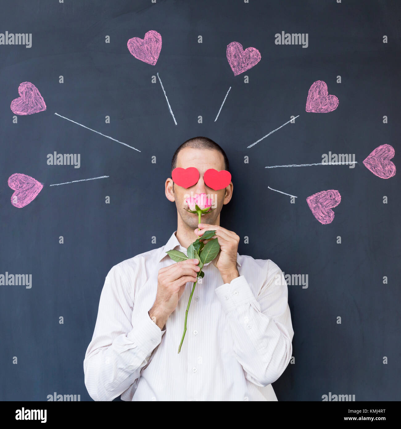 Singolo adulto uomo bianco con gli occhi a forma di cuore che indossa una camicia bianca e in piedi di fronte a una lavagna con dipinto di cuori tenendo una rosa. Foto Stock