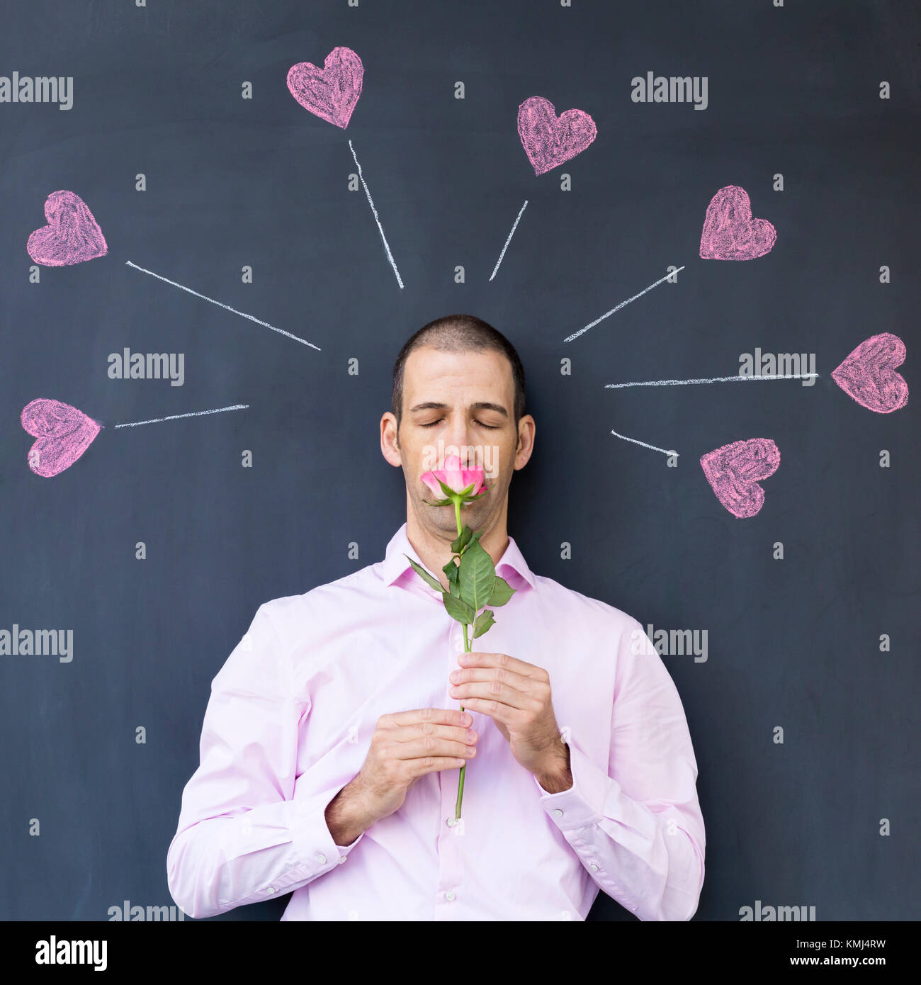 Singolo adulto uomo bianco che indossa una camicia rosa in piedi di fronte a una lavagna con dipinto di cuori tenendo una rosa. Concetto di amore folle. Foto Stock