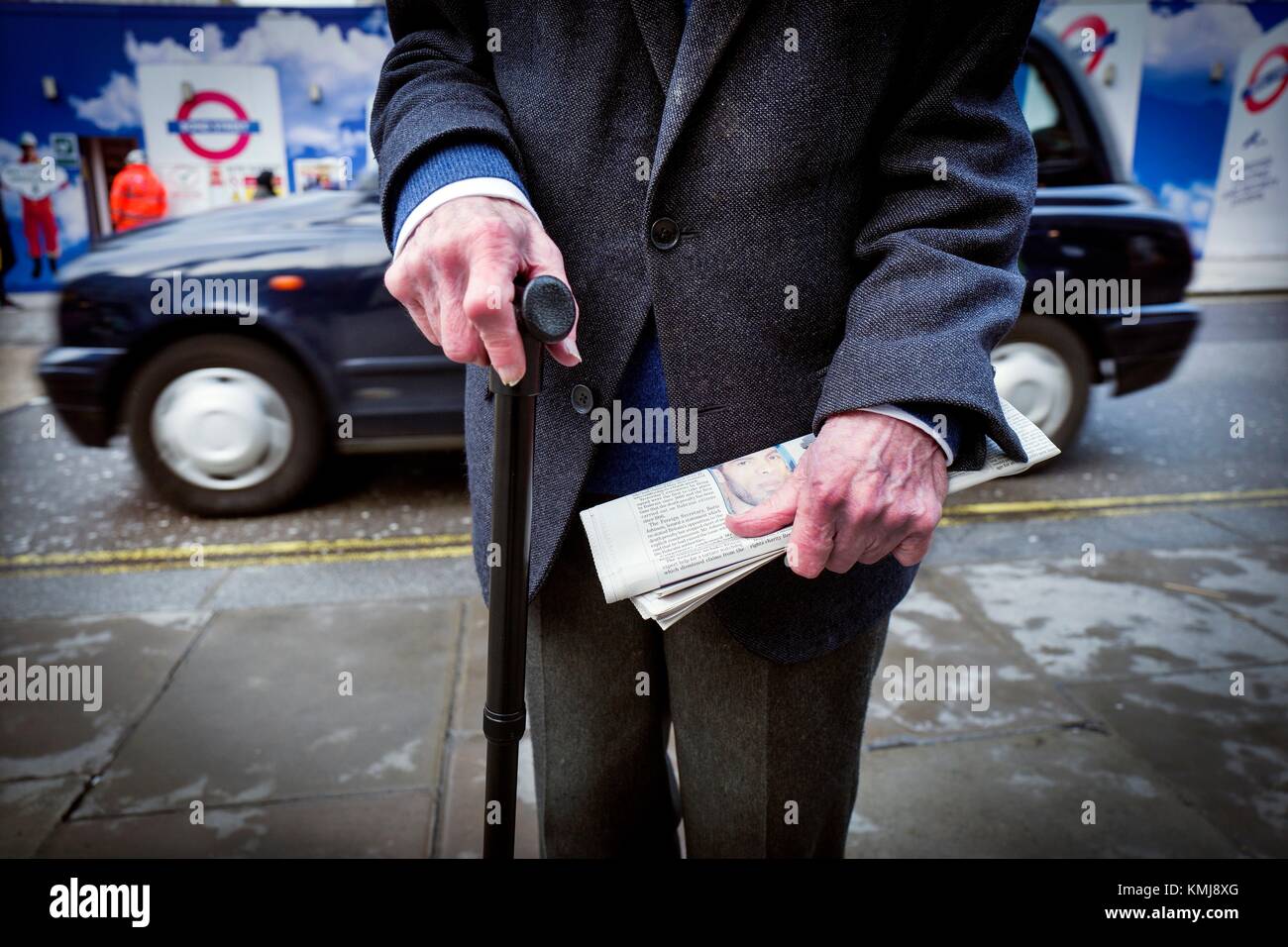 Il Primer plano de onu hombre mayor irreconocible con onu baston en una mano y onu periodico en la otra, al fondo ONU taxi. Oxford Street, Londres, Regno Unito, Europa. Foto Stock