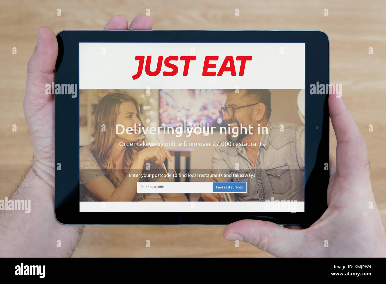 Un uomo guarda al solo mangiare sito sul suo iPad dispositivo tablet, sparato contro un tavolo di legno top sfondo (solo uso editoriale) Foto Stock
