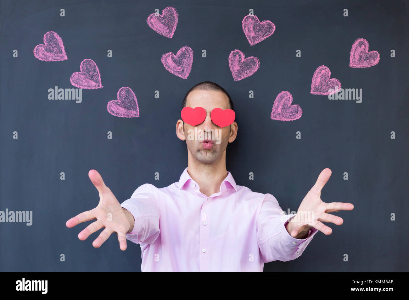 Singolo adulto uomo bianco con gli occhi a forma di cuore che indossa una camicia rosa e in piedi di fronte a una lavagna con dipinto di cuori si distende le sue mani. Foto Stock