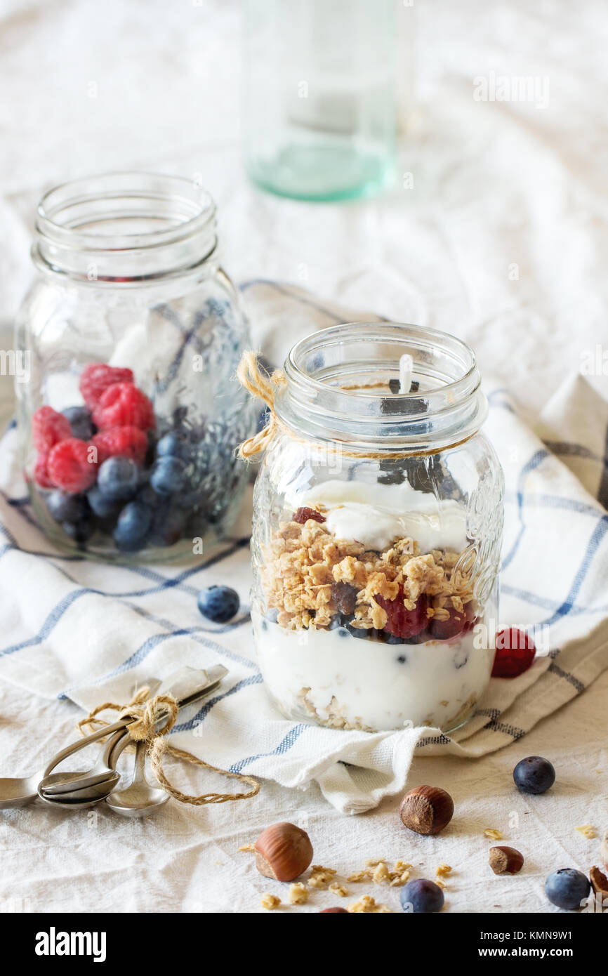 Vaso di vetro con muesli fatte in casa e yogurt servito con dadi, lamponi e more su bianco tessile in luce del giorno Foto Stock