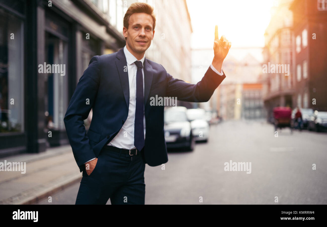 Sorridente giovane imprenditore in un vestito in piedi su una strada di Città al mattino con il suo braccio in aria salutando un taxi Foto Stock