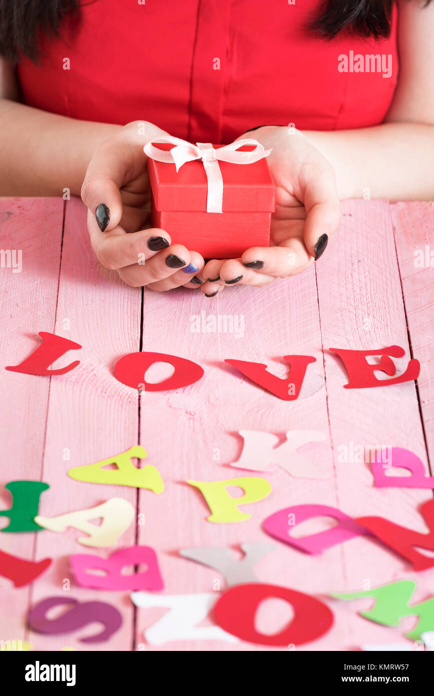 Donna mani tenendo una confezione regalo rossa su una rosa tavolo in legno e piena di carta multicolore di lettere e la parola amore scritto fuori di essi. Foto Stock