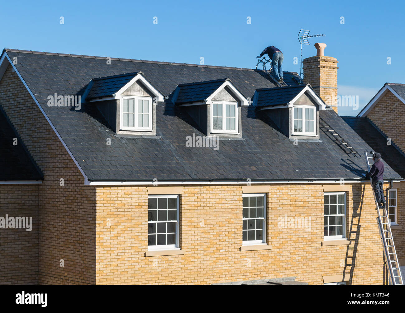 Le riparazioni di prendere posto per un tetto di ardesia su una casa di mattoni con tre finestre dormer. Gli uomini conciatetti sono su scale di alta. Un uomo è sulla parte superiore del tetto. Ther Foto Stock