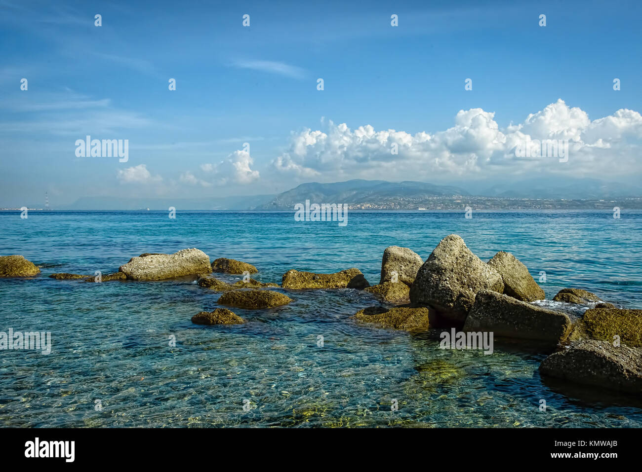 Il mare limpido e cristallino con rocce in primo piano sotto un cielo azzurro con soffici nuvole bianche Foto Stock