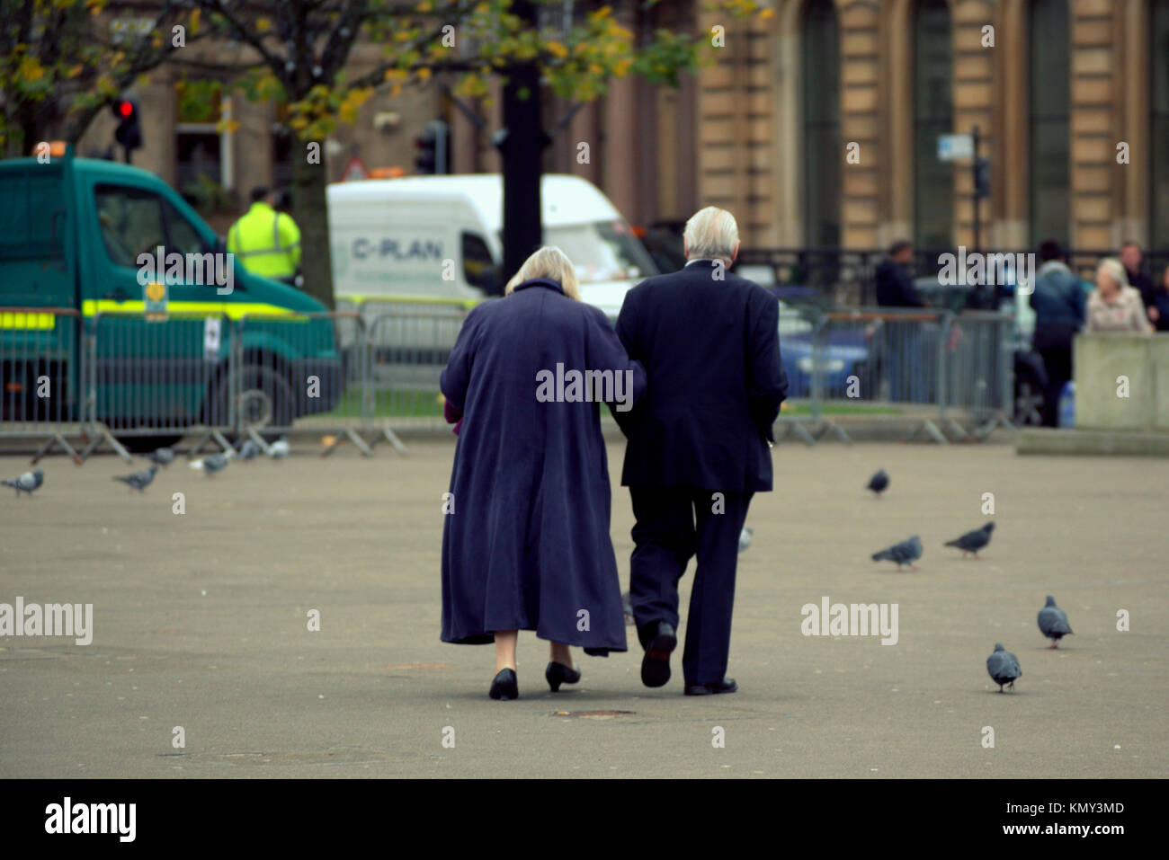 Vecchia coppia di anziani uomo e la moglie donna che indossa blu navy attraversando a piedi piazza tra i piccioni visto da dietro George Square,Glasgow, Regno Unito Foto Stock