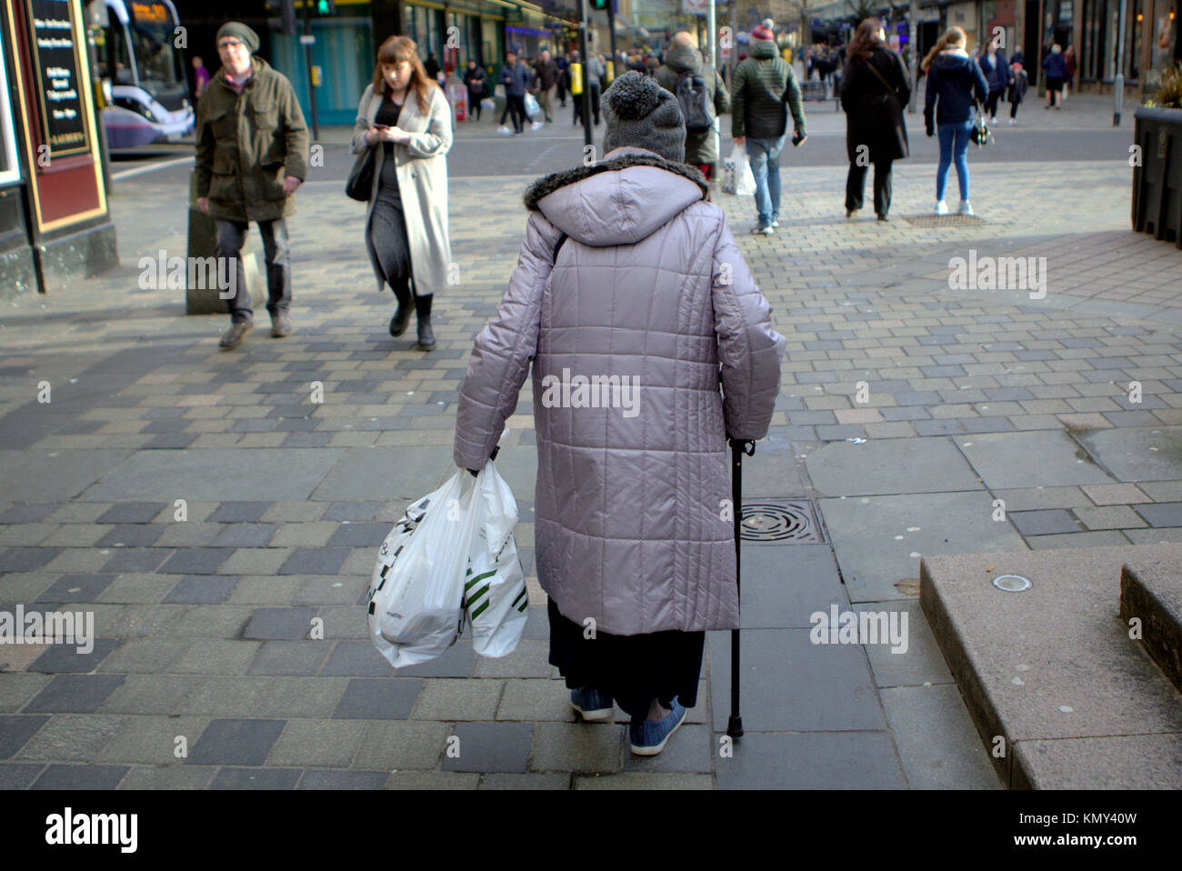 Old Lady senior citizen alle prese con un bastone da passeggio e shopping bags visto da dietro sulla Sauchiehall Street zona pedonale di Glasgow Foto Stock