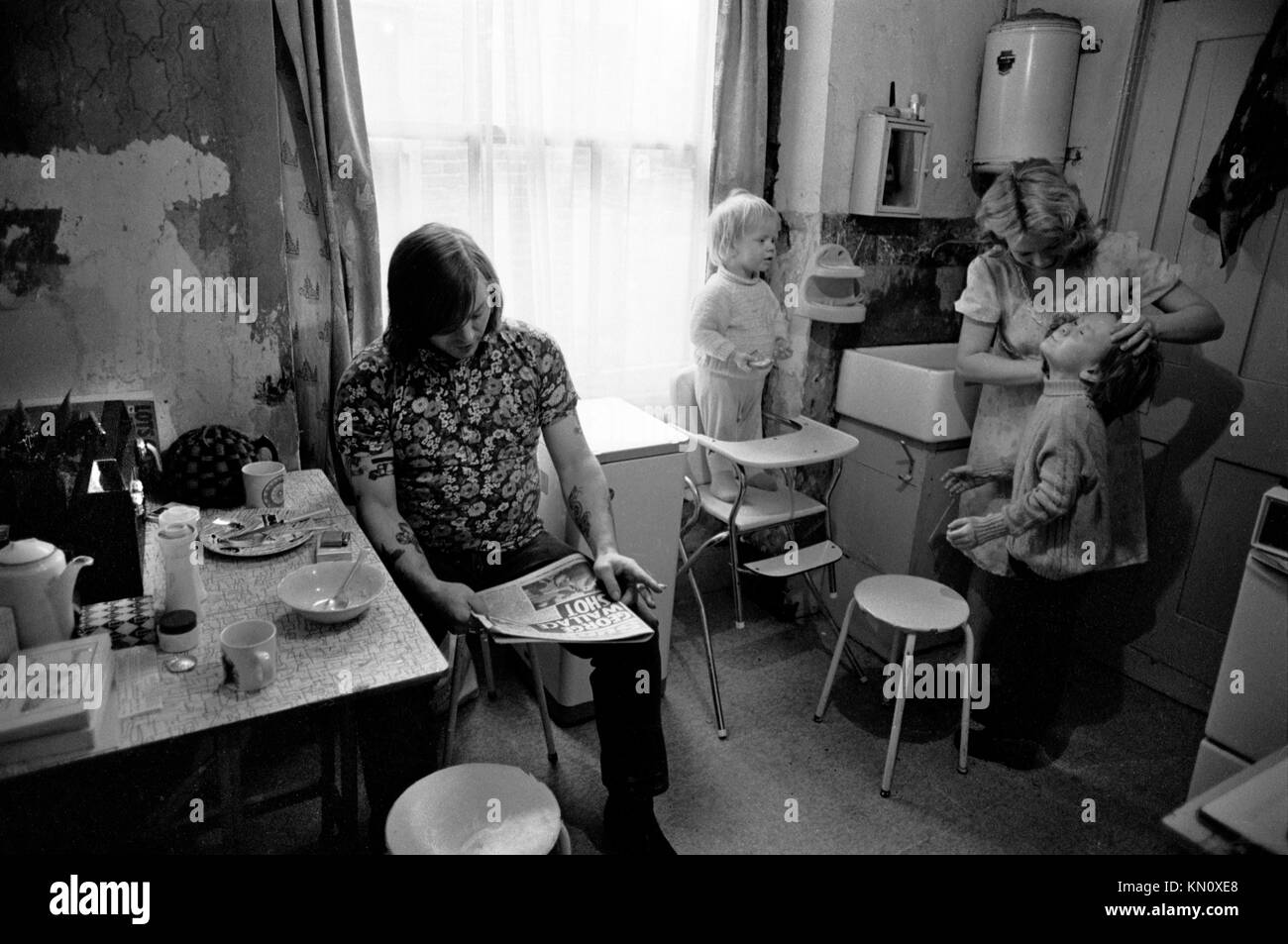 1970s Slum Housing, cucina doppia come bagno. Famiglia che mangia insieme. Povertà. Scaldabagno a gas Ascot sulla parete sopra il lavandino del maggiordomo 1972 Fulham, Inghilterra anni '70 UK HOMER SYKES Foto Stock