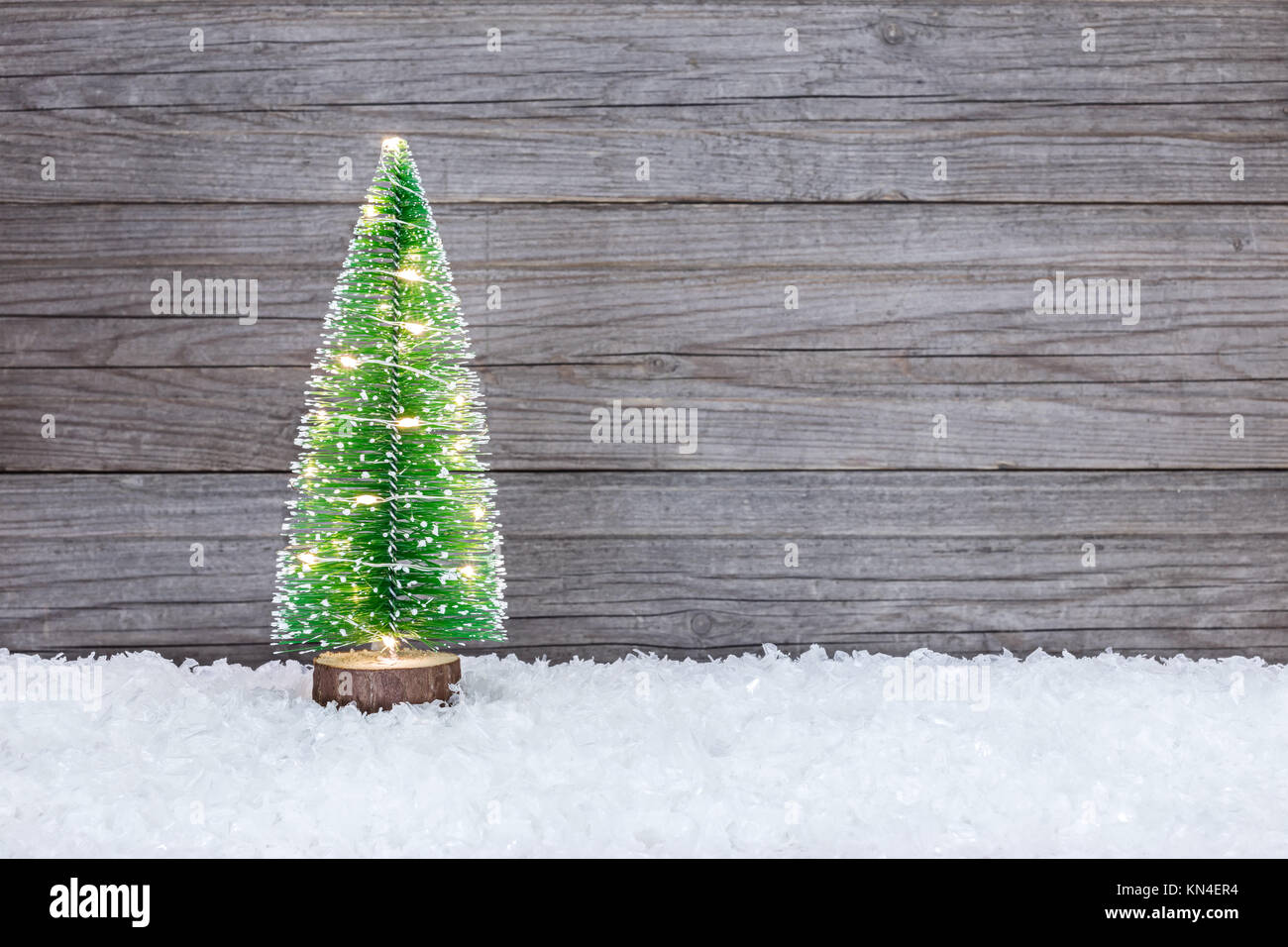 Piccola verde artificiale albero di Natale illuminato con luci delle vacanze sulla neve contro di legno sfondo rustico Foto Stock