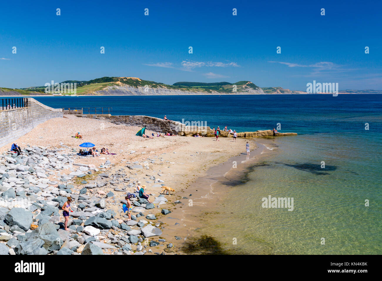 I villeggianti godendo il sole in spiaggia a est, Lyme Regis con cappuccio dorato al di là su Jurassic Coast Sito Patrimonio Mondiale, Dorset, England, Regno Unito Foto Stock