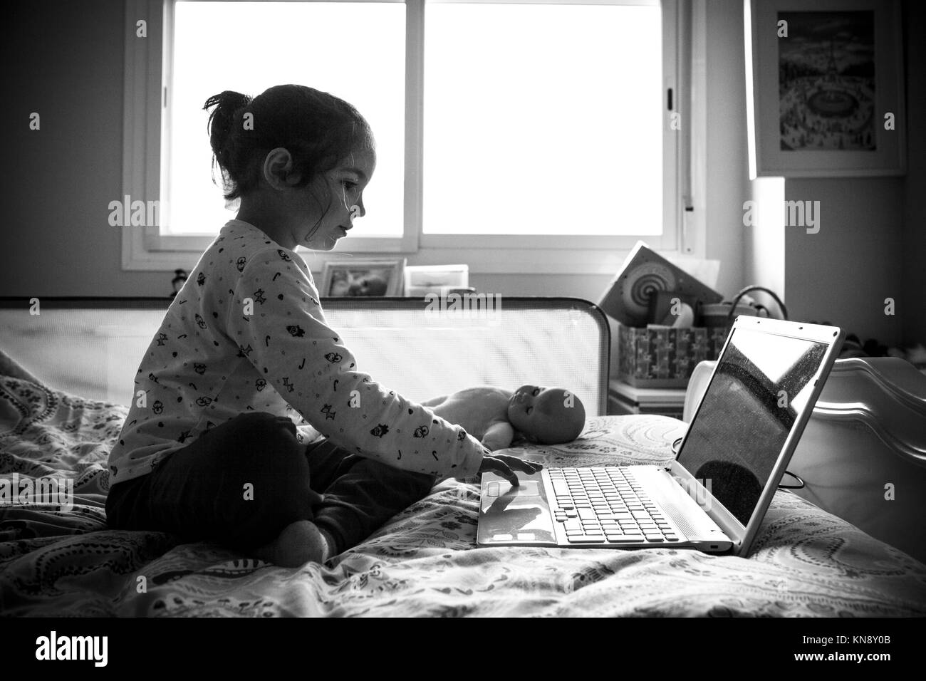 Poco ragazza seduta nel letto e giocare ai videogame on line nella sua camera da letto. In bianco e nero girato. Foto Stock
