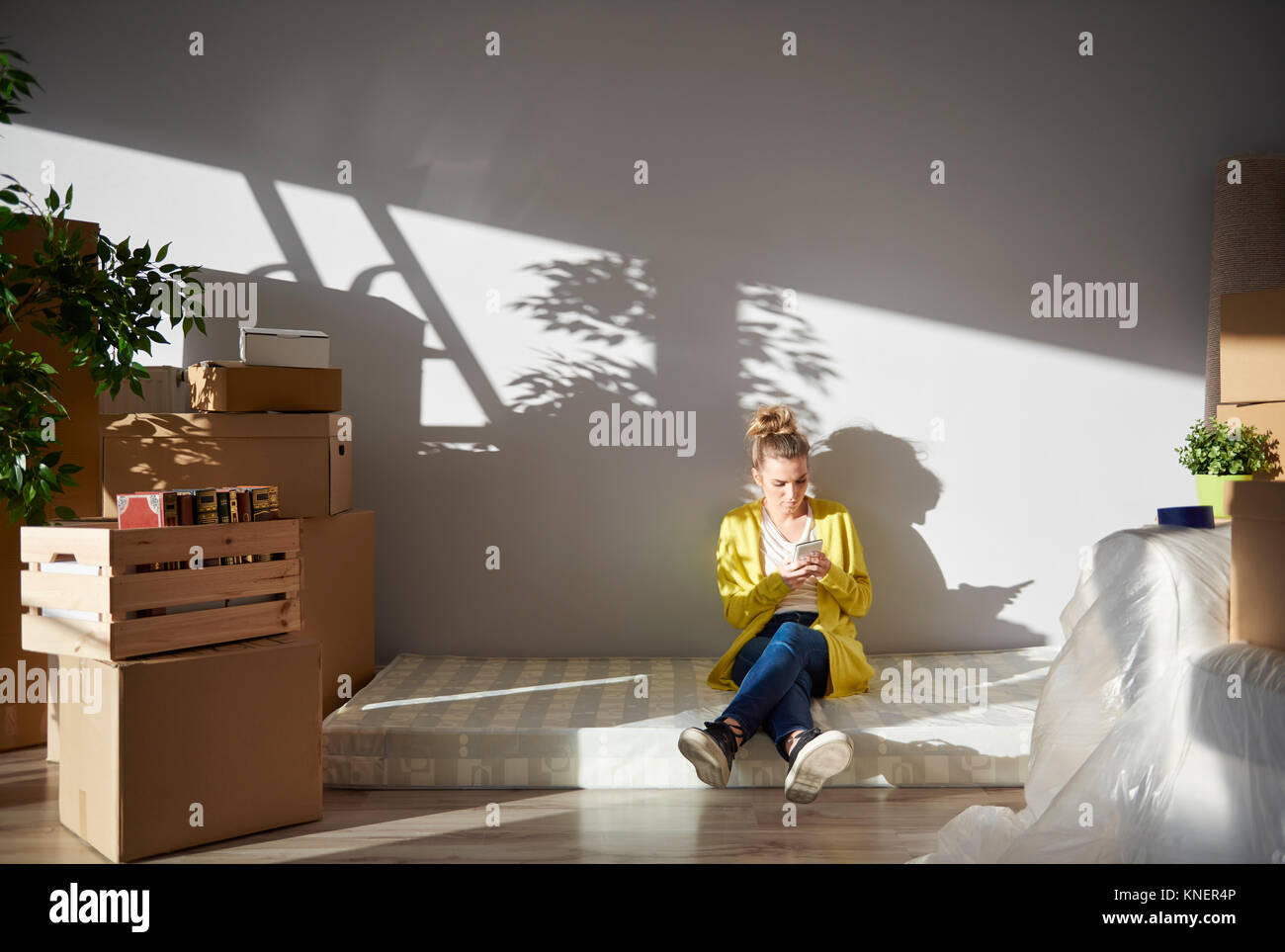 Giovane donna seduta sul materasso nella nuova casa, utilizza lo smartphone Foto Stock