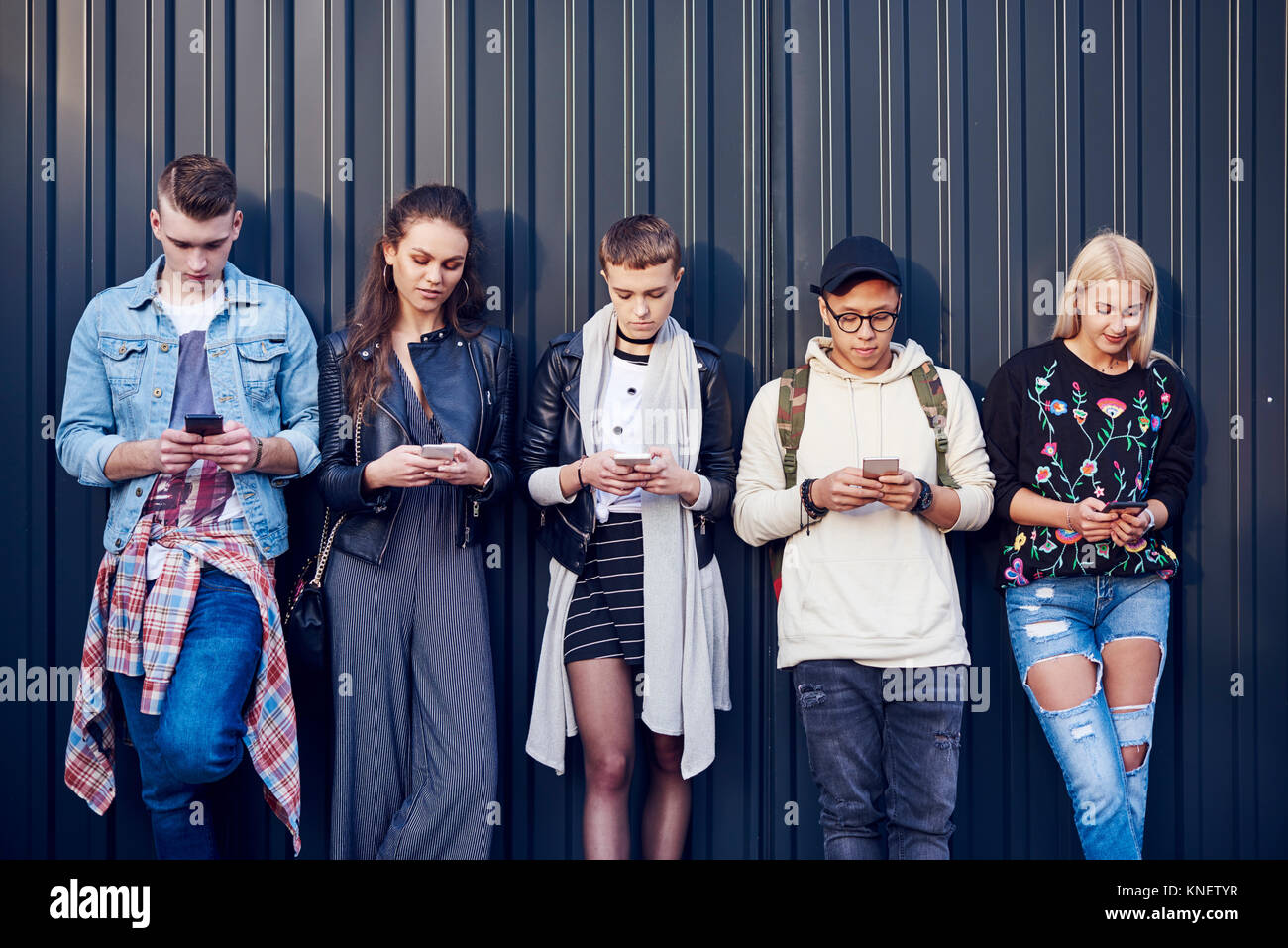 Fila di cinque giovani amici adulti appoggiata contro la parete nera cercando smartphone a Foto Stock