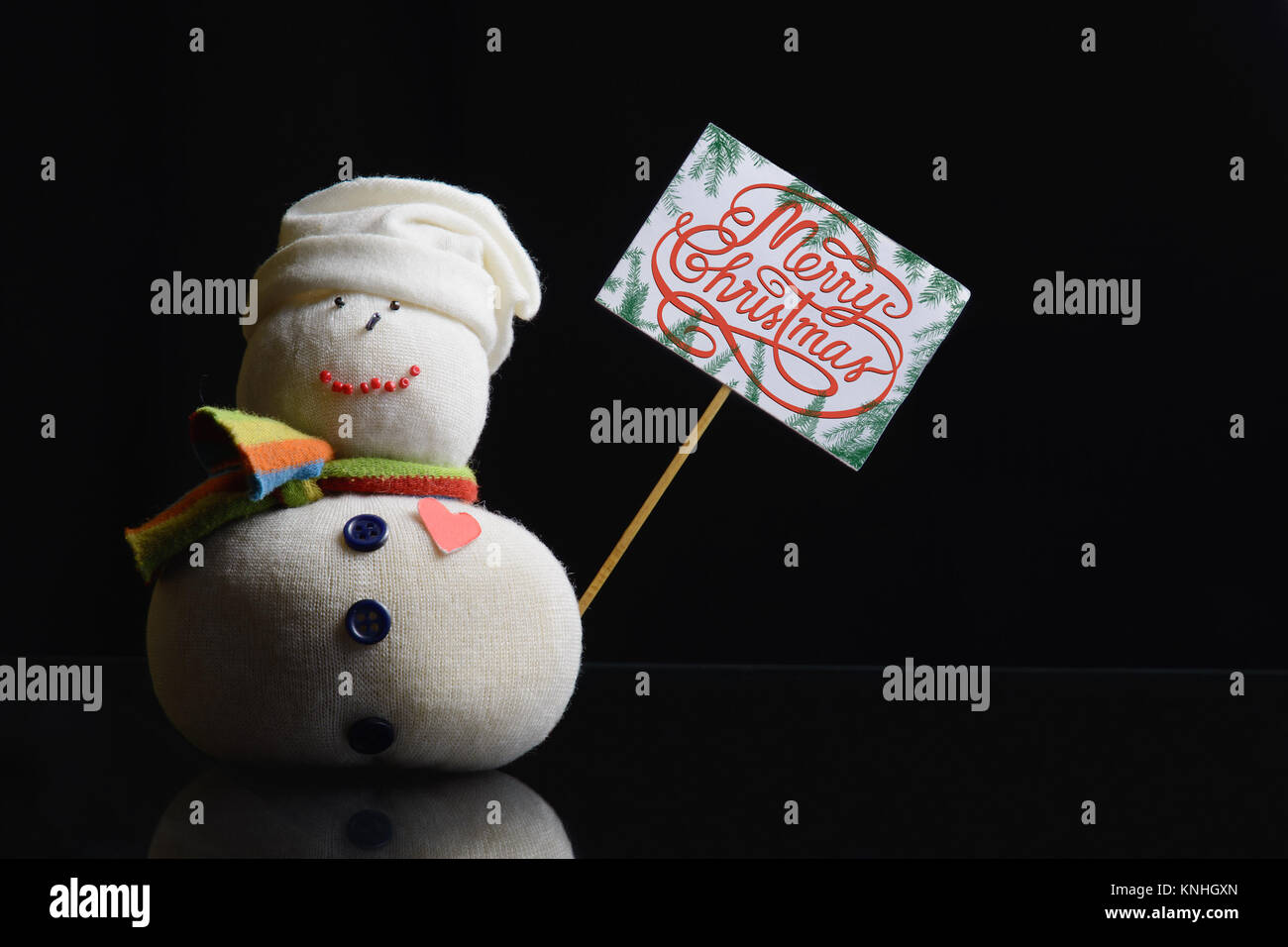 Pupazzo di neve figura tenendo un cartello di cartone su un bastone con il testo "Buon Natale!" scritto su di esso, e circondato con rami di abete. Tasto basso fotografia. Foto Stock