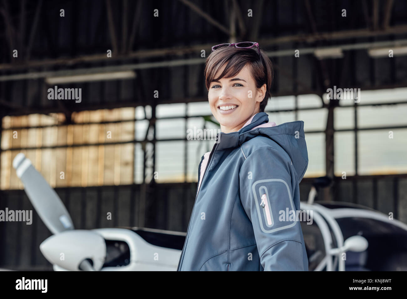 Allegro sorridente giovane donna in posa l'aeroporto hangar con il suo piccolo velivolo, viaggi e concetto di aviazione Foto Stock