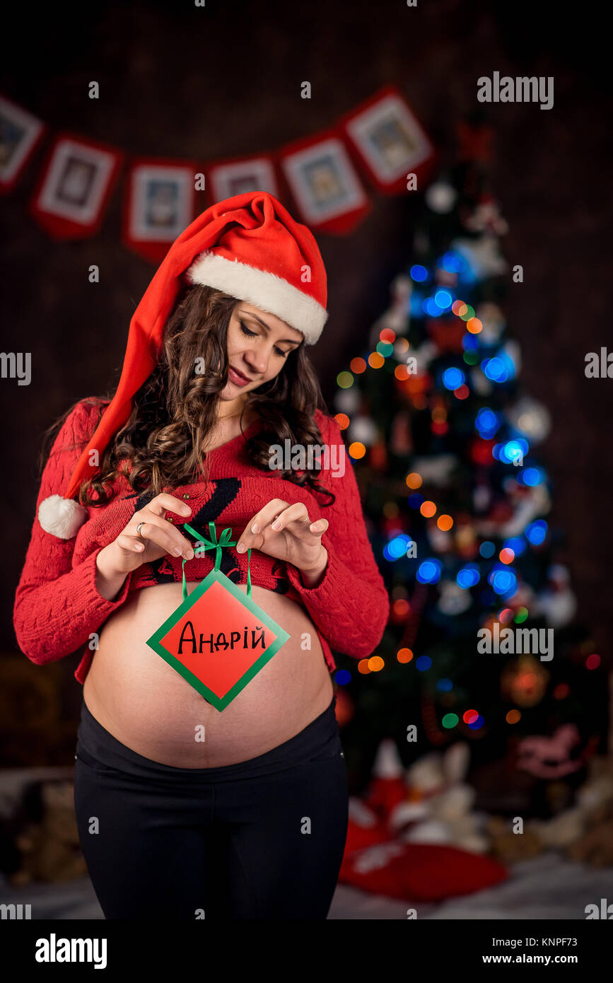 Immagini Di Natale Donne.A Mezza Lunghezza Ritratto Della Bella Donna Incinta Con Capelli Ricci Che Indossa Il Berretto Di Babbo Natale E Tenendo Premuto Il Segno Andrii Sullo Sfondo Di Un Albero Di Natale Foto
