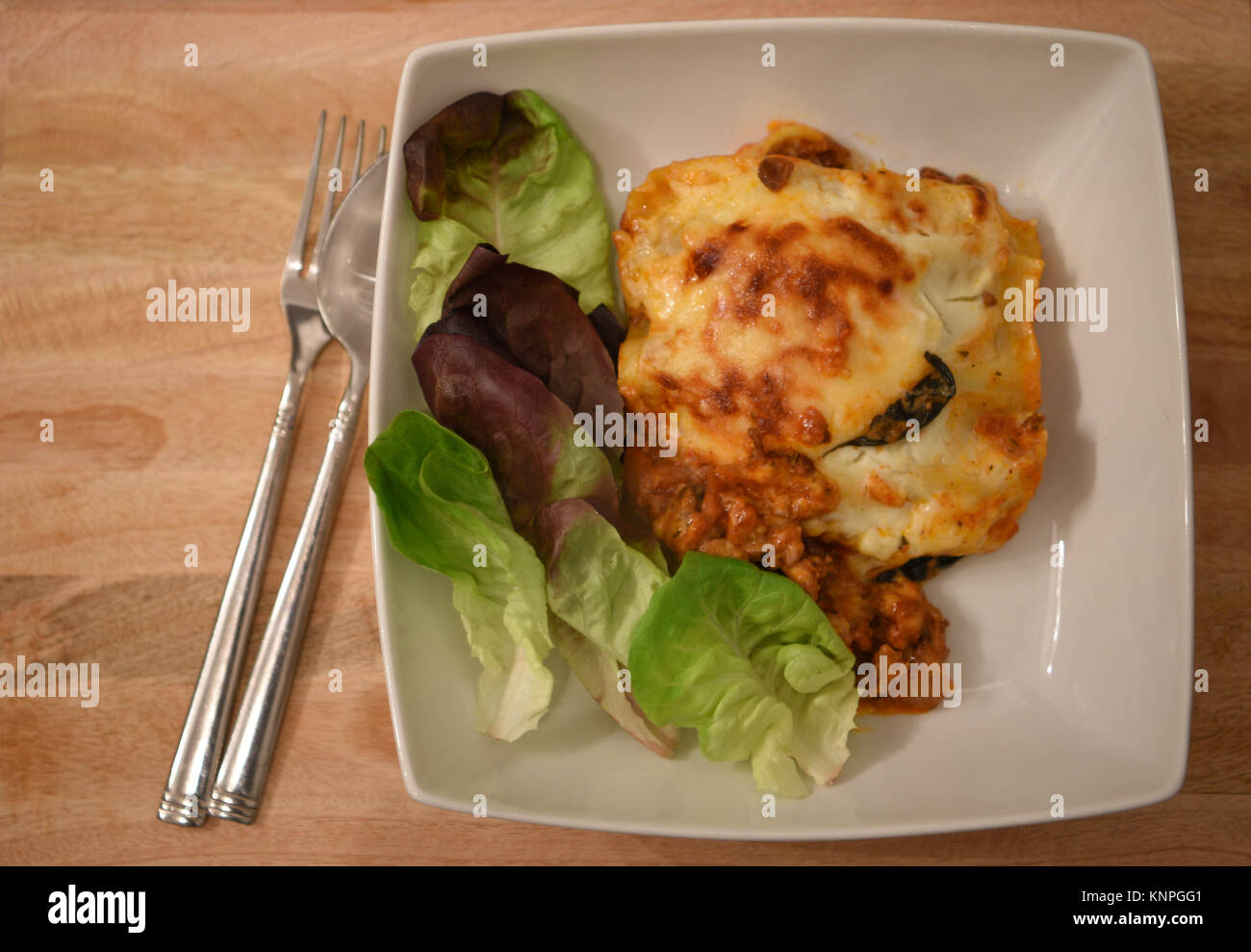 Cibo fotografia immagine di una porzione di una deliziosa casa calda cuocere le lasagne con foglia verde insalata in piatto bianco con posate e un background in legno Foto Stock