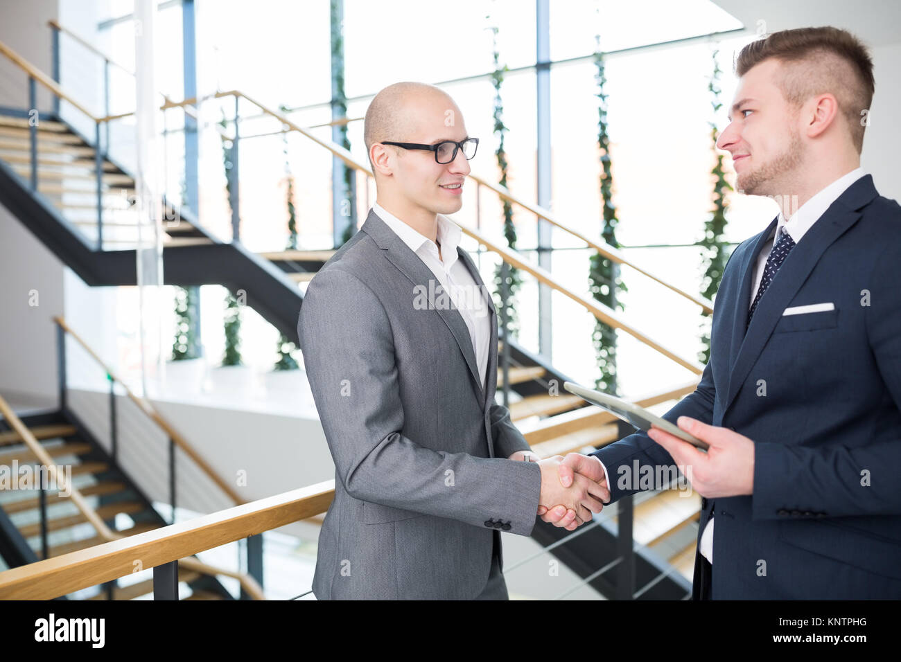 Fiducioso imprenditore tenendo tavoletta digitale mentre si stringono le mani con il collega di sesso maschile contro la scala in ufficio Foto Stock