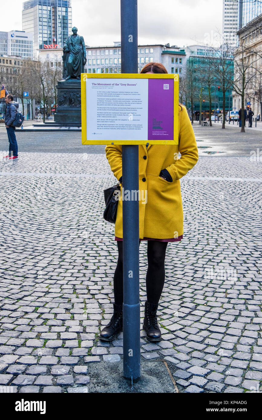 Francoforte, Germania.giovane donna in giallo cappotto grigio legge gli autobus monumento information board. Memorial commemora l assassinio di disabili e malati Foto Stock