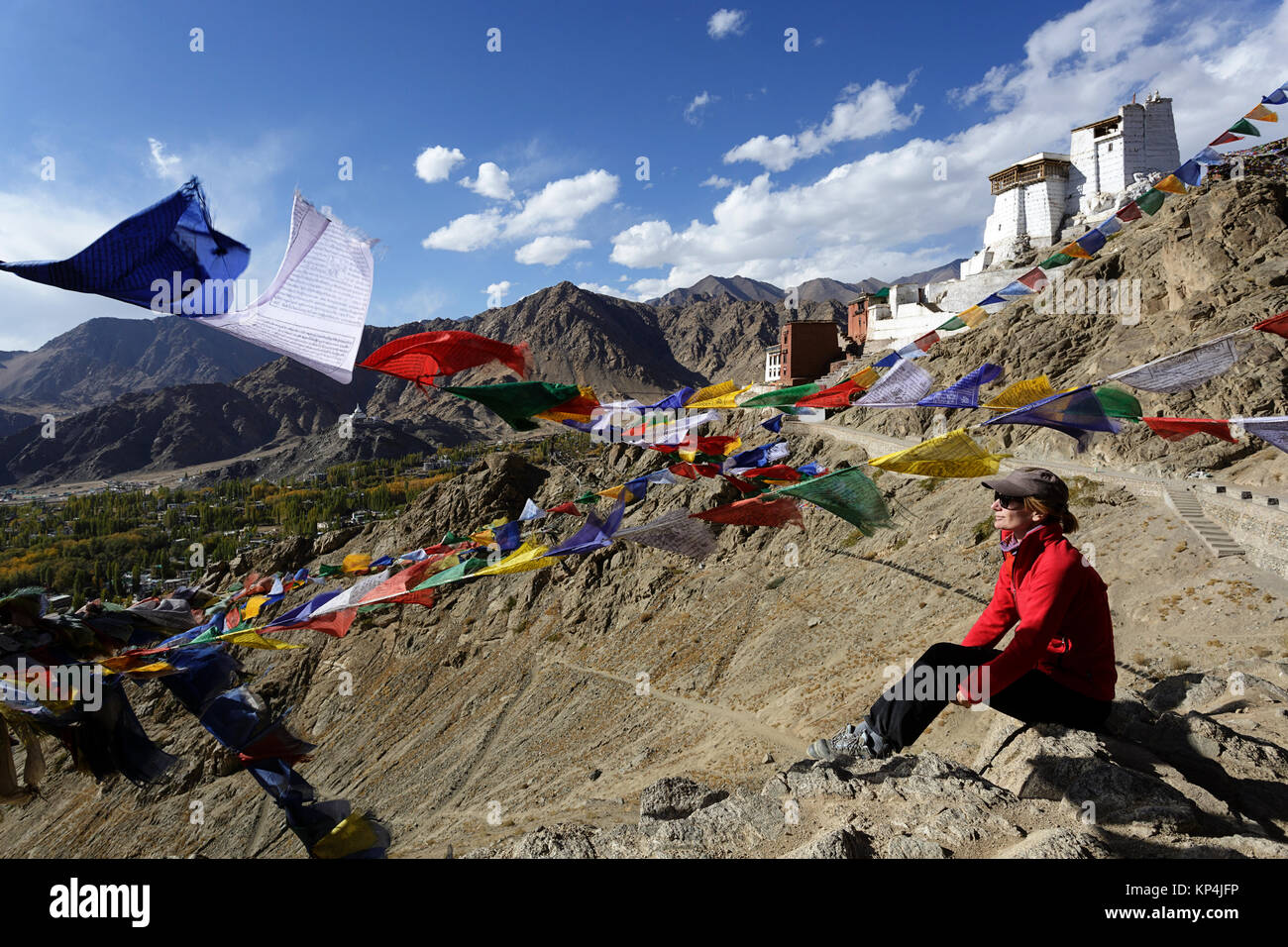 Woman in Red Jacket seduto su di una roccia sotto le bandiere di preghiera con Tsemo Gompa in background, Leh, Ladakh, Jammu e Kashmir in India. Foto Stock