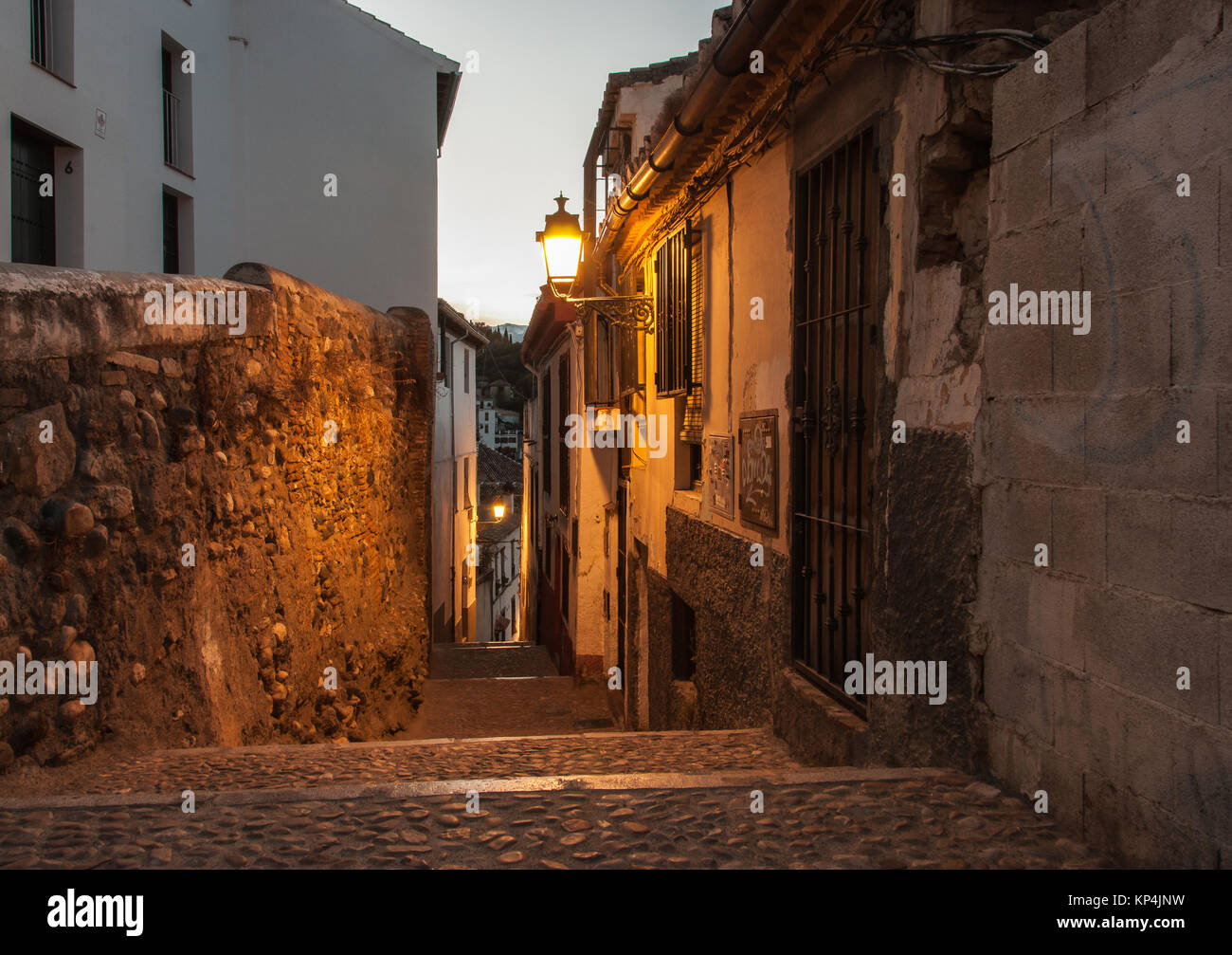 Un ripide e strette strade in ciottoli di una città medievale nella calda luce dei lampioni contrastano con il bianco plasterd pareti e pallido cielo mattutino. Foto Stock