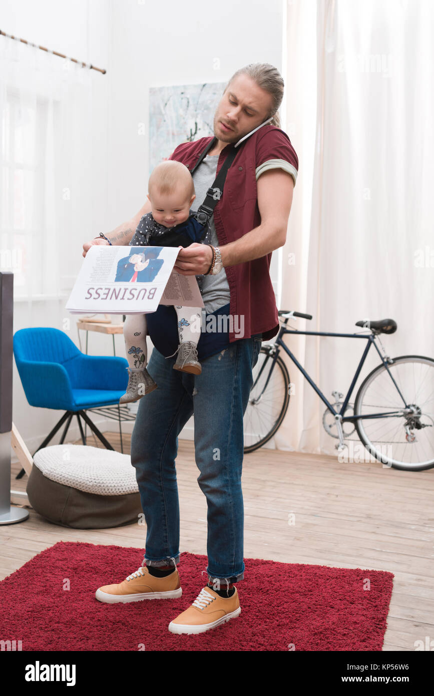 Padre in piedi con il figlio in baby sling, tenendo il giornale e parlare tramite smartphone Foto Stock
