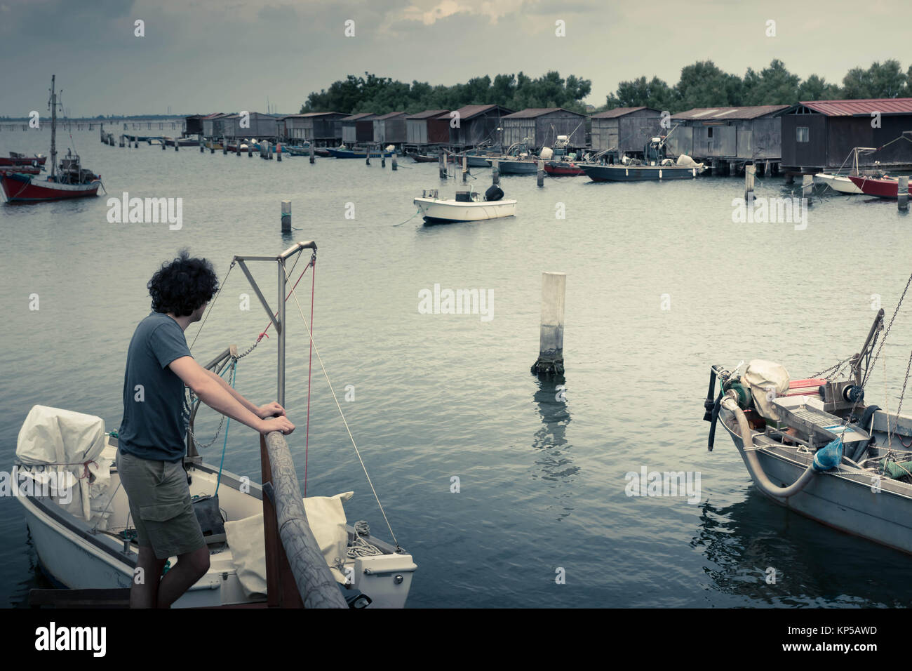 Dissaturato lago paesaggio con un ragazzo su un molo in legno a guardare le barche dei pescatori e la pesca capannoni. Parco del Delta del Po laguna. Veneto, Italia. Foto Stock