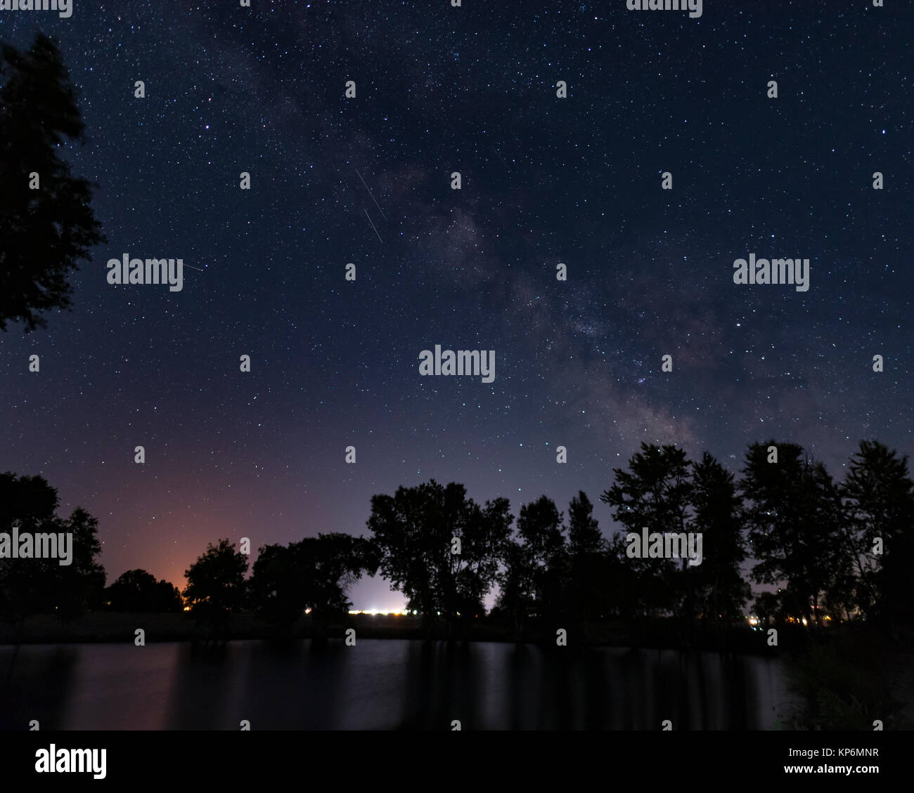 Scena notturna con la Via Lattea sopra le acque scure di un laghetto con alberi in Sihouette Foto Stock