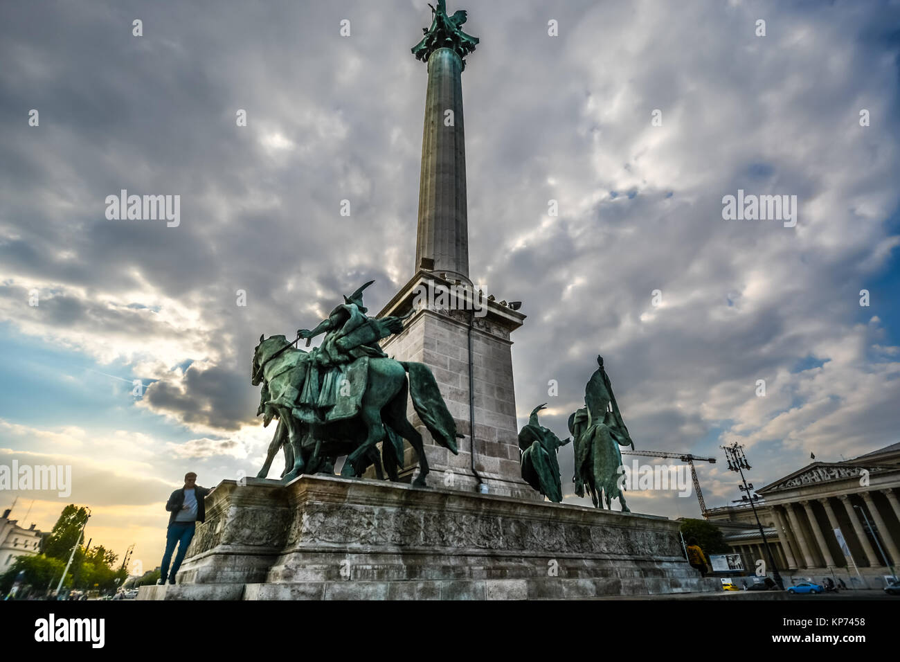 Un giovane uomo cammina sul Millenium monumento in Piazza degli Eroi, o Hosok tere presso il parco della città di Budapest in Ungheria su un pomeriggio nuvoloso Foto Stock