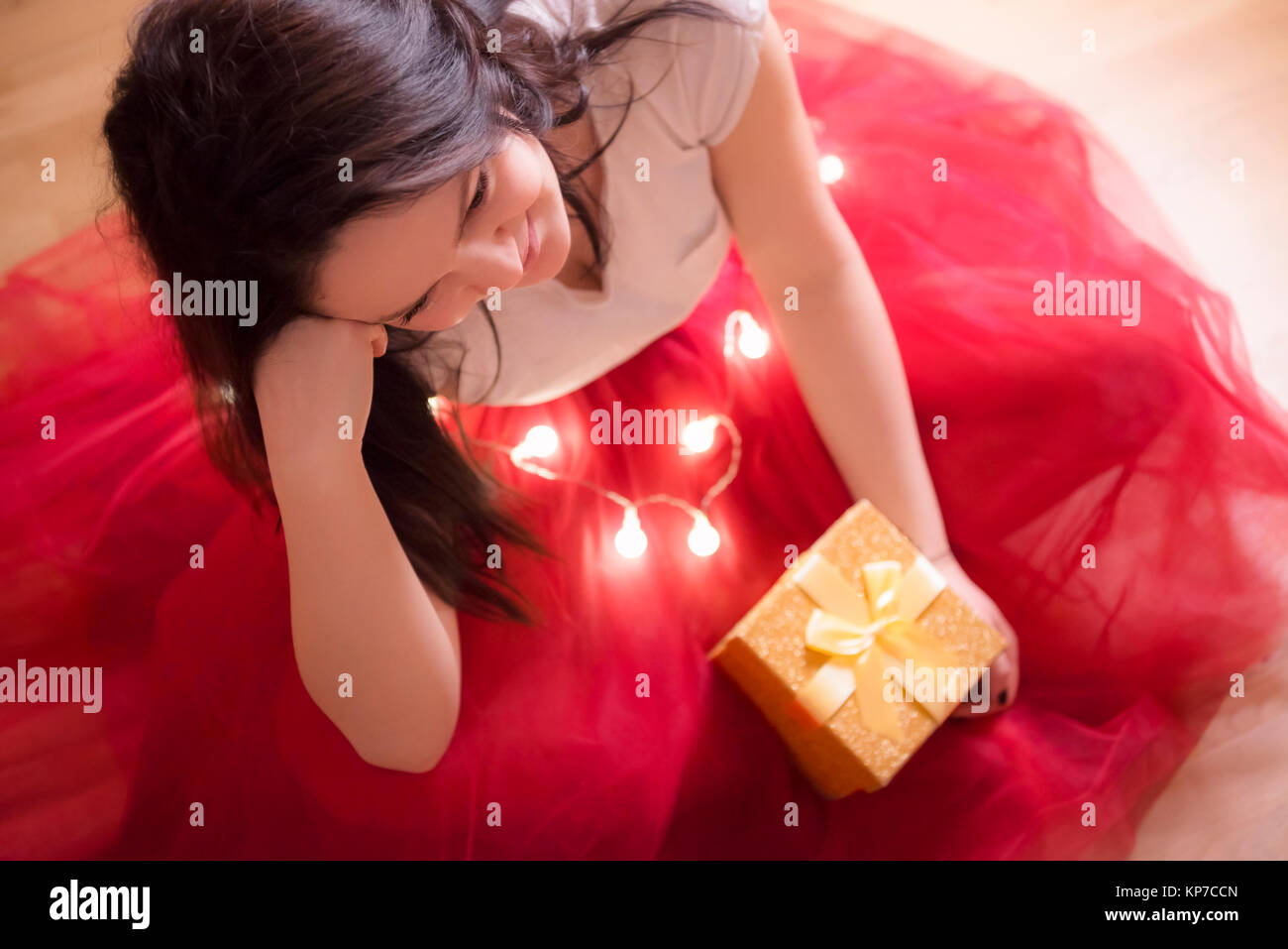 Giovane e bella bruna donna vestita di un rosso tutu, seduto sul pavimento, circondato da luci di Natale e in possesso di una confezione regalo con parità di prua. Foto Stock
