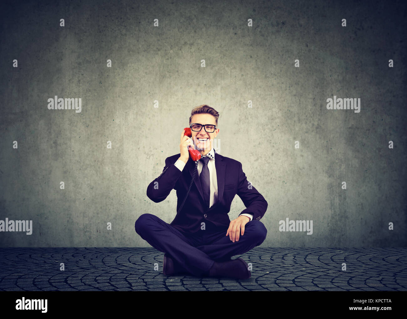 Uomo in tuta seduta con le gambe incrociate e parlando al telefono allegramente contro uno sfondo grigio. Foto Stock