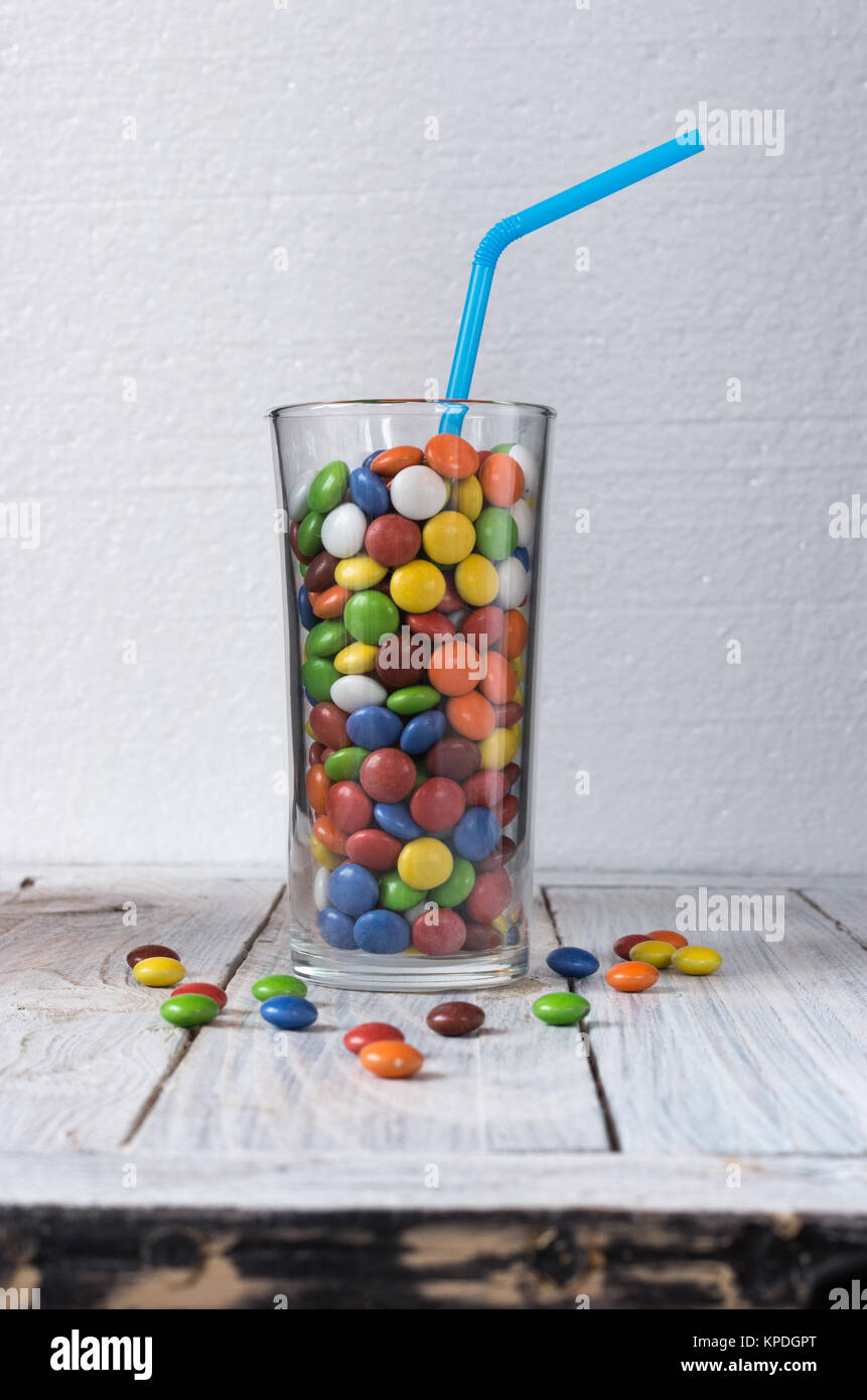 Il vetro è riempita con caramelle colorate. Un bicchiere con un drink e una cannuccia. Vista frontale. Caramelle sono sparsi sul tavolo. Foto Stock