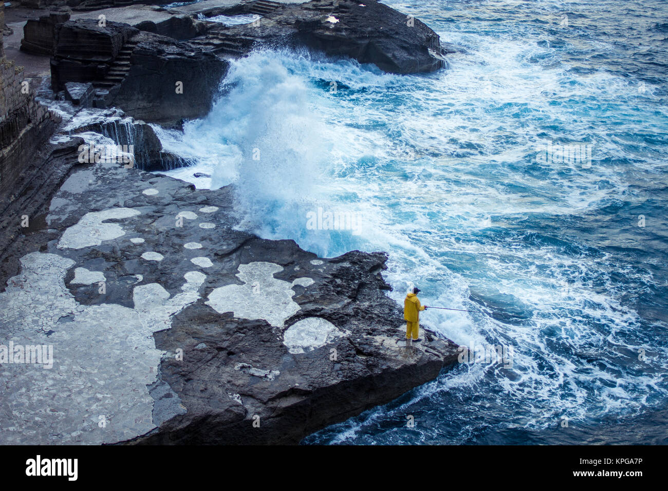 Un pescatore solitario in piedi sul bordo di una roccia di fronte a una massiccia ondata di colpire la riva Foto Stock