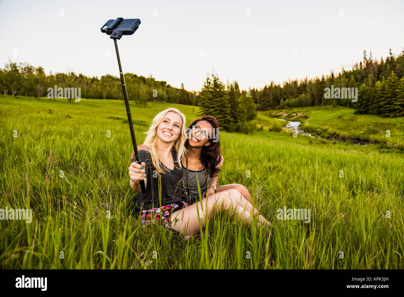 Due amiche seduto in un campo di erba in posa per un autoritratto con un bastone selfie e smart phone; Edmonton, Alberta, Canada Foto Stock