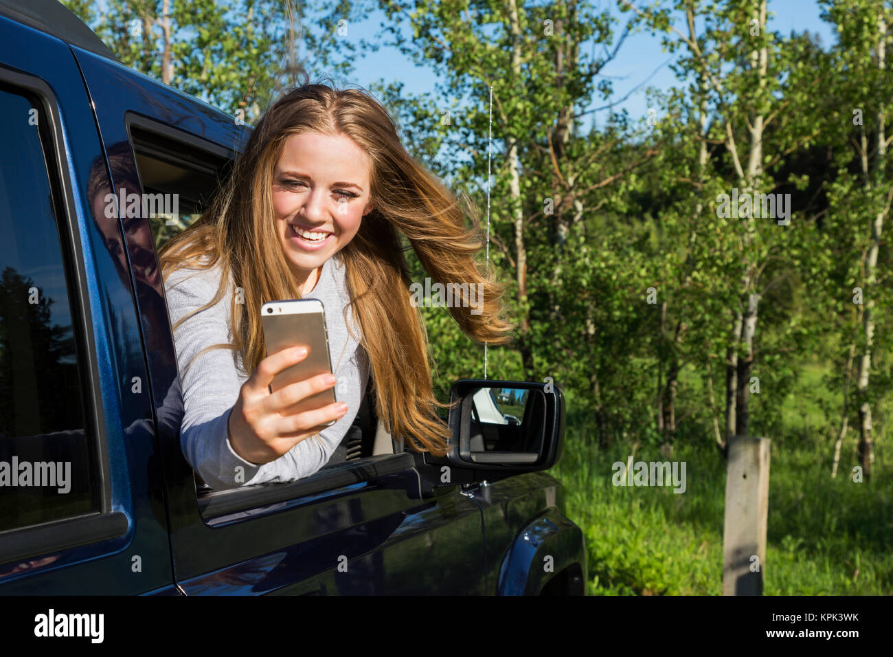 Una giovane e bella donna con capelli lunghi biondi guardando fuori un cristallo di un veicolo e prendendo un autoritratto con il suo smart phone; Edmonton, Alberta, Canada Foto Stock
