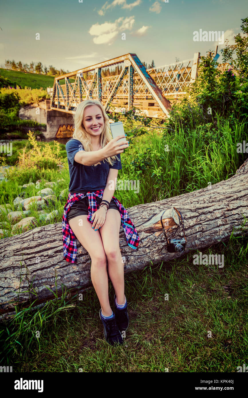 Una giovane donna siede su un log in un parco prendendo un autoritratto con il suo telefono cellulare con un ponte e un fiume in background; Edmonton, Alberta, Canada Foto Stock
