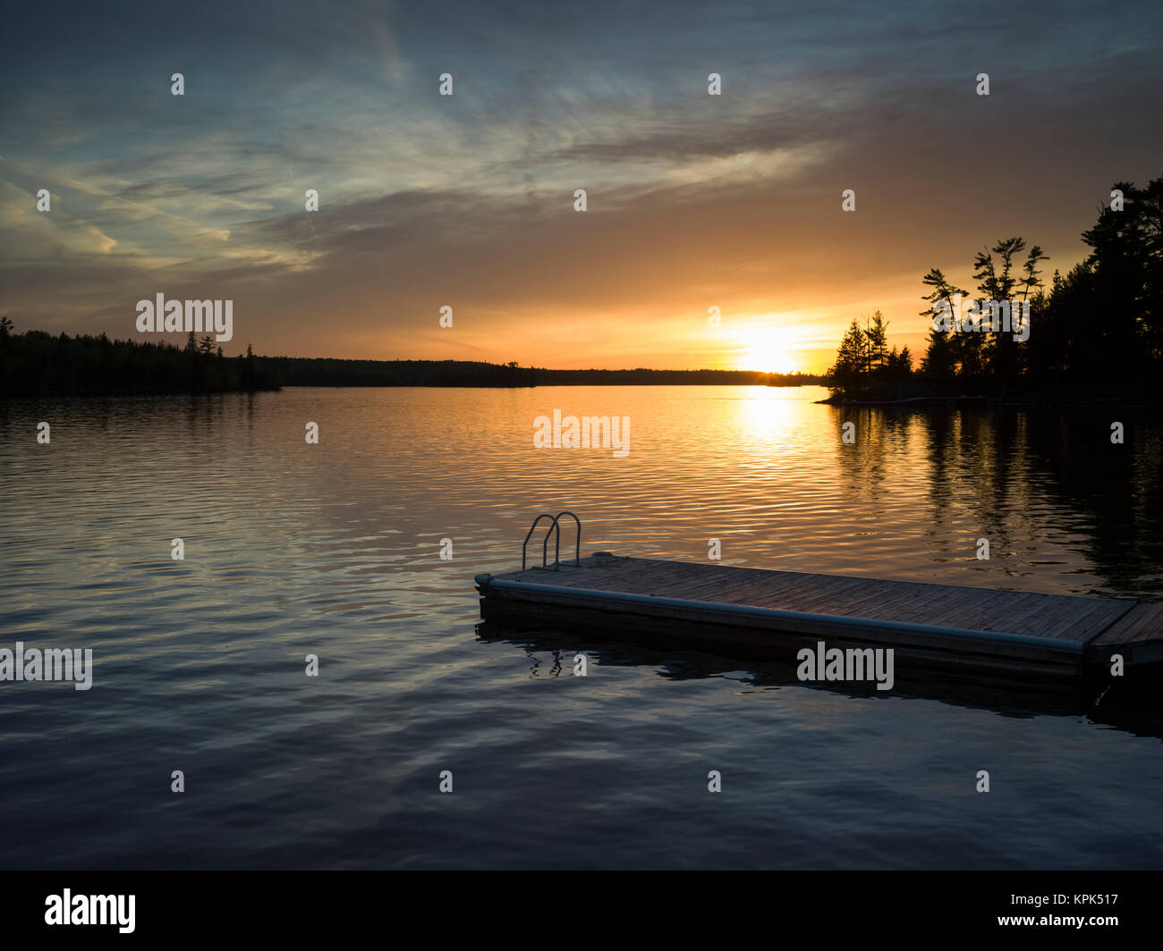 Il sole che tramonta su un tranquillo lago e stagliano alberi lungo il litorale con un dock e la scaletta in primo piano Foto Stock