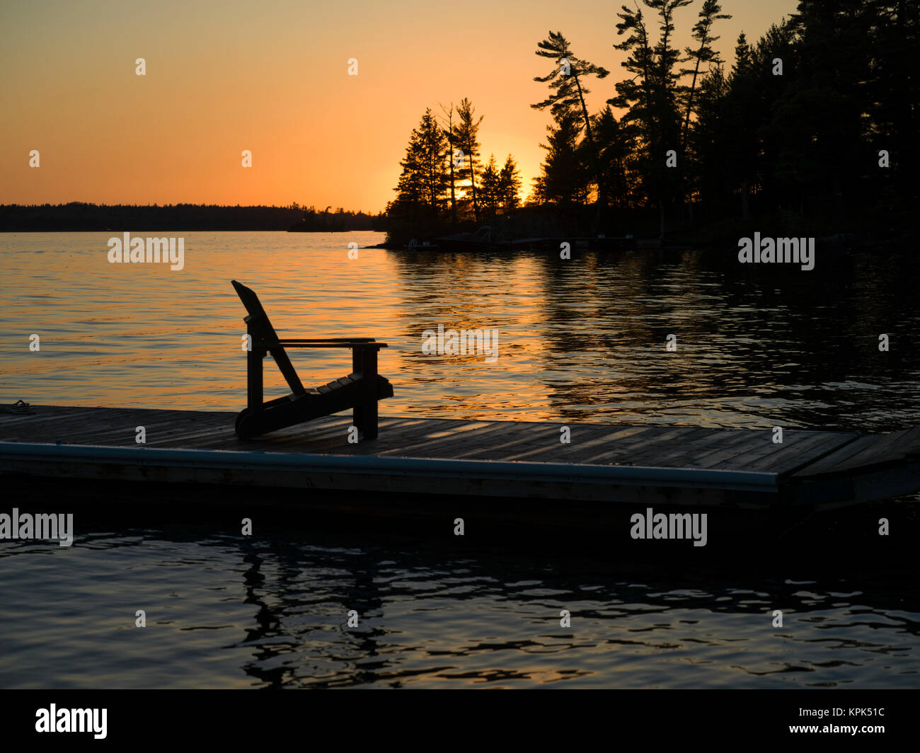 Un legno sedia Adirondack siede su un dock su un tranquillo lago al tramonto; lago dei boschi, Ontario, Canada Foto Stock