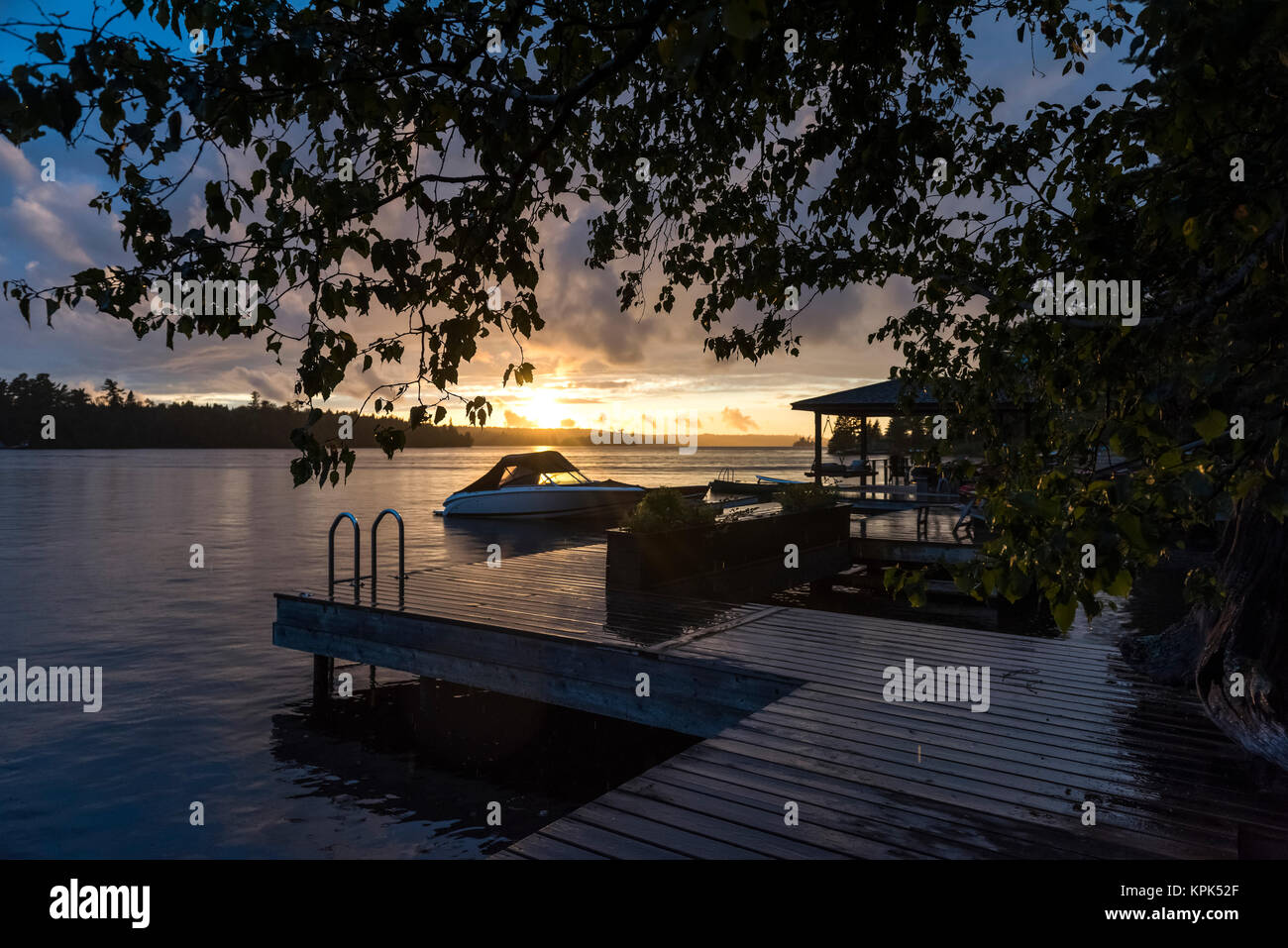 Il sole che tramonta su un tranquillo lago con un dock e la barca in primo piano; il lago dei boschi, Ontario, Canada Foto Stock