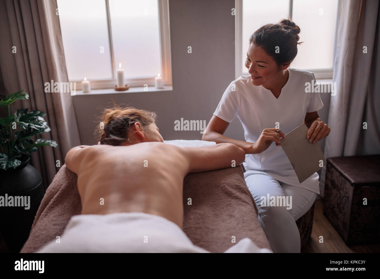 Estetista professionista a parlare con la femmina sdraiato sul lettino per massaggi. Massaggiatrice asiatica parlando con client femmina prima del trattamento termale wellness cen Foto Stock