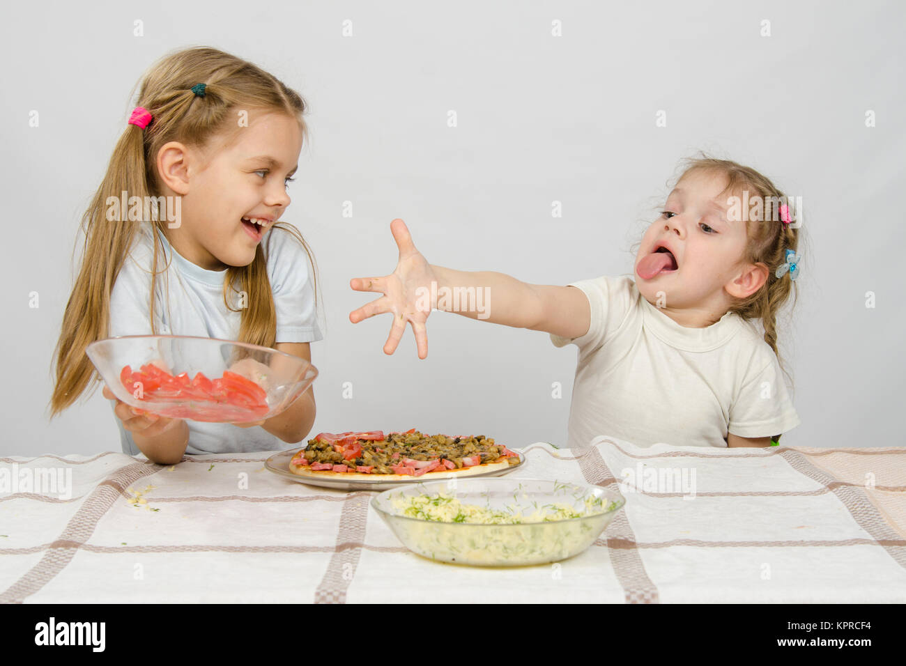 Due bambine in una tabella preparata la pizza. Uno con una bizzarra vista si allunga una mano per una piastra con pomodori, che prende gli altri con un sorriso Foto Stock