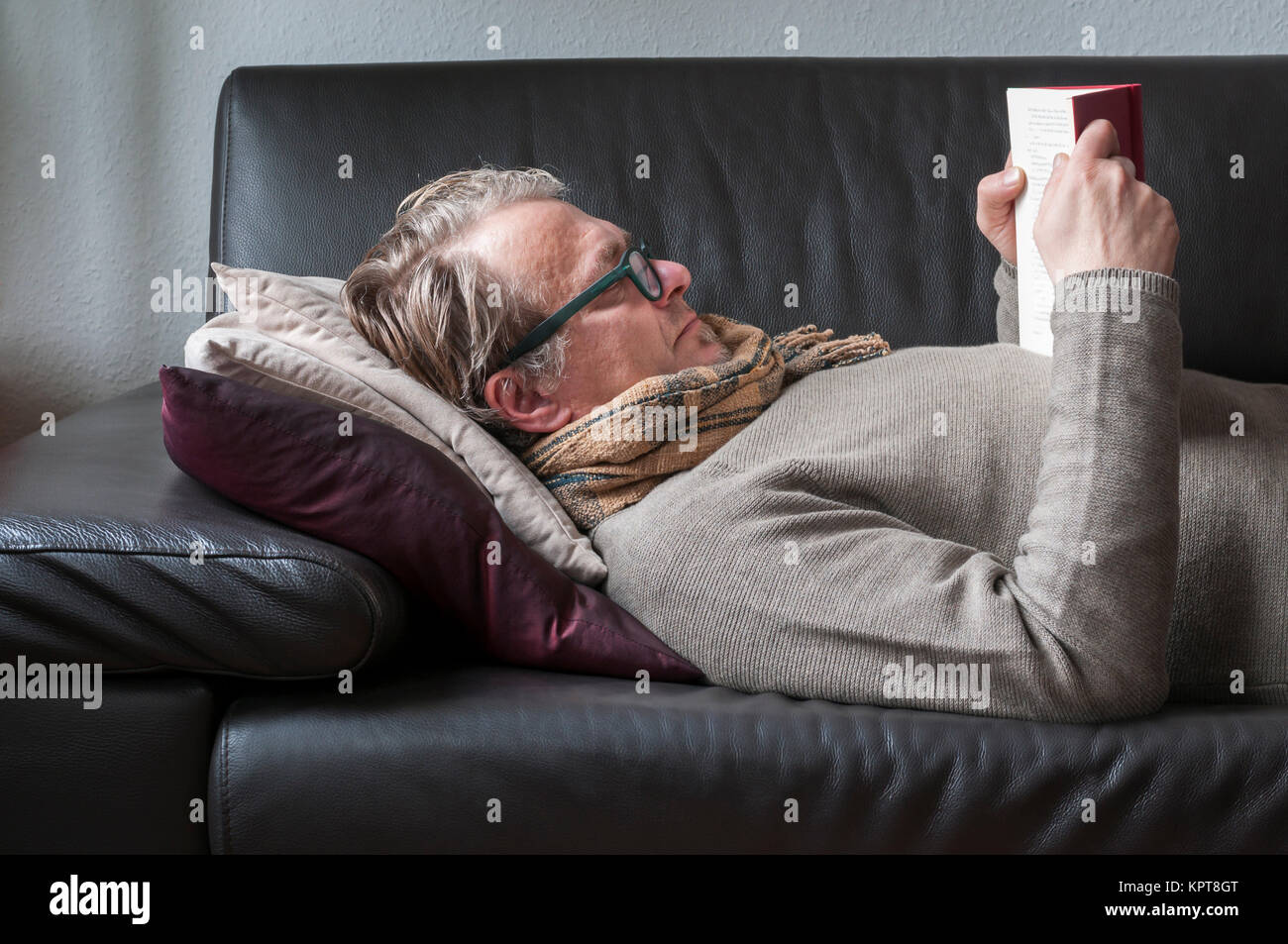 Liegender Mann auf einem divano mit Brille und Schal liest in einem Buch. Foto Stock