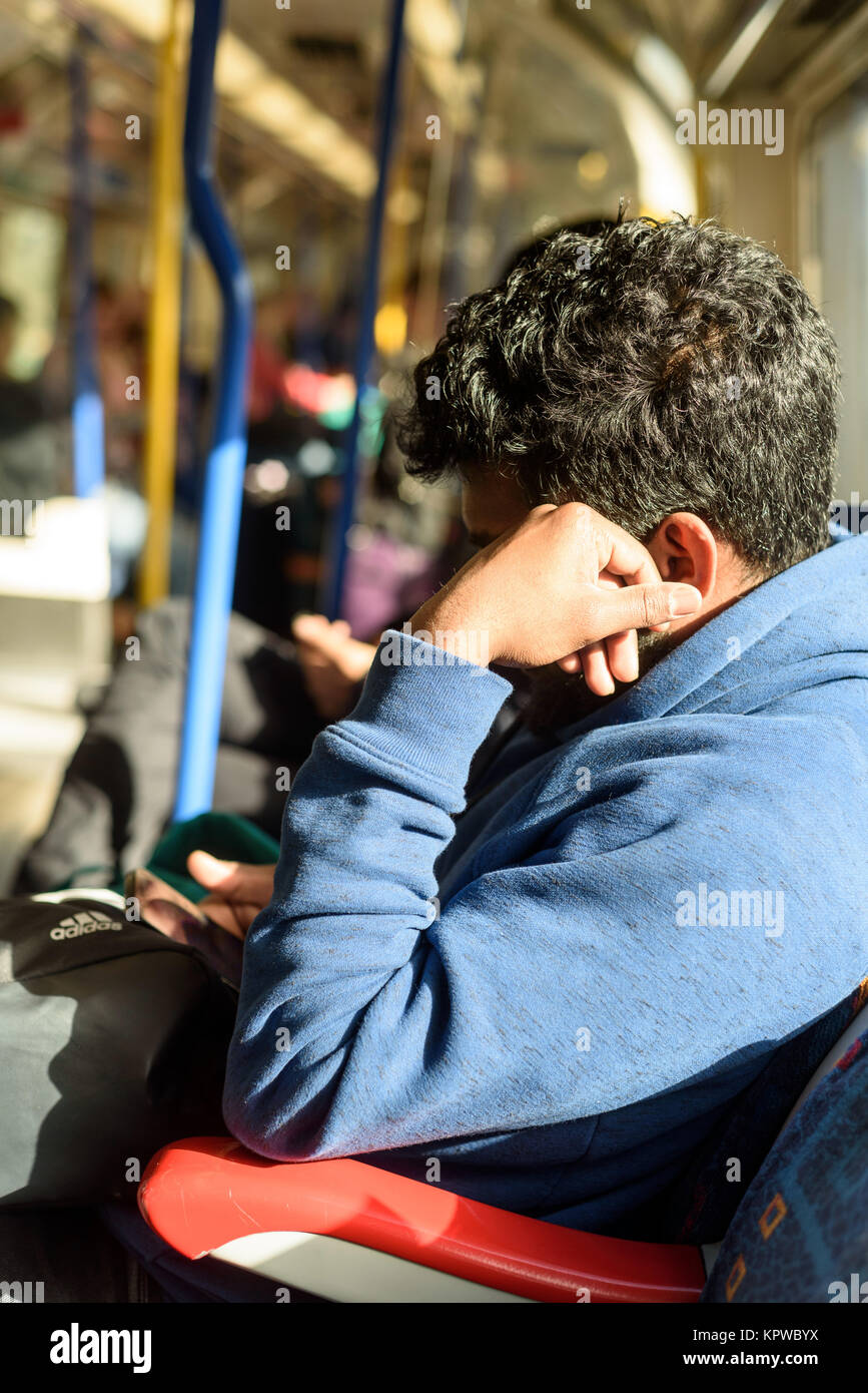 Asian Indian uomo con un telefono nella luce solare naturale usando il suo smartphone mentre in sella alla London metropolitana linea di treno Foto Stock