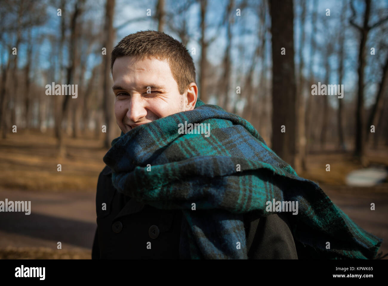 Giovane uomo è godendo il sole e sorridente in un gelido inverno mattina in piedi nel parco vicino agli alberi. Una persona Foto Stock